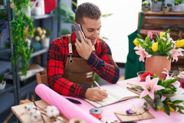 Young caucasian man florist talking on smartphone writing on notebook at flower shop