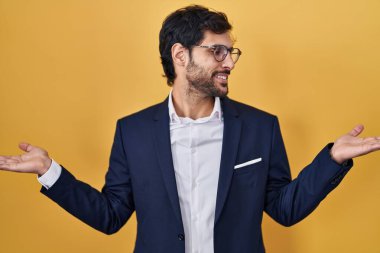 Handsome latin man standing over yellow background smiling showing both hands open palms, presenting and advertising comparison and balance 