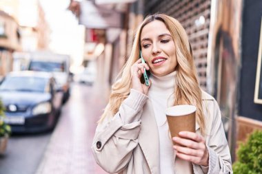 Young blonde woman talking on smartphone drinking coffee at street