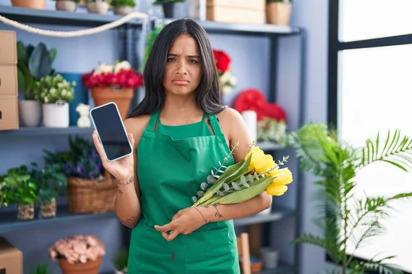 Brunette woman working at florist shop holding smartphone skeptic and nervous, frowning upset because of problem. negative person.