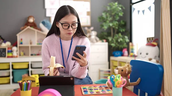 Young Chinese Woman Preschool Teacher Eating Banana Using Smartphone Kindergarten — Stock fotografie