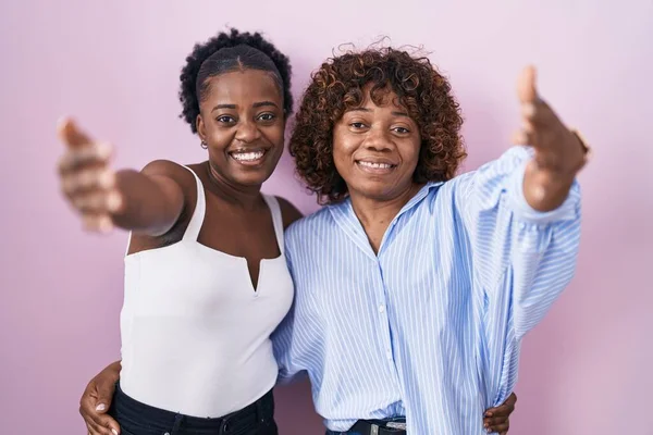 ピンクの背景の上に立っている2人のアフリカの女性は 抱擁のために開いて腕で笑顔カメラを見ている 幸せを受け入れる陽気な表情 — ストック写真