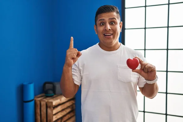 Hispanic young man holding red heart at gym smiling with an idea or question pointing finger with happy face, number one