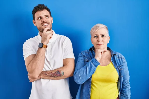 青い背景の上に立っている若いブラジル人の母親と息子は 質問を心配し 顎に手で心配し 緊張しています — ストック写真