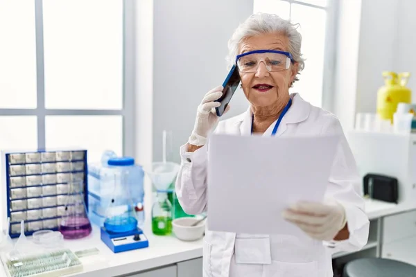 研究室でスマートフォンで話す科学者の制服を着たシニア白髪の女性 — ストック写真