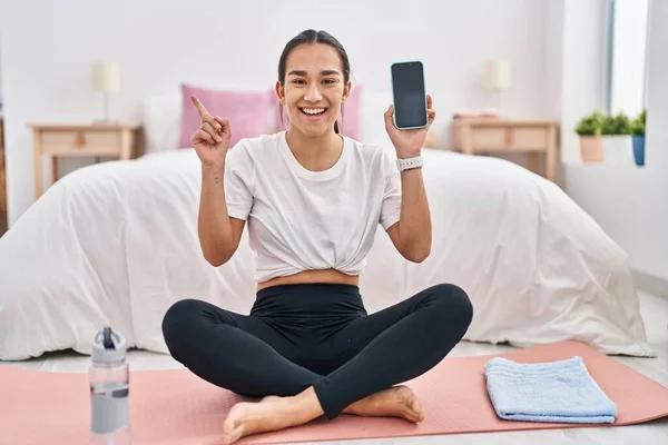 Young south asian woman doing yoga mat holding smartphone showing screen smiling happy pointing with hand and finger to the side