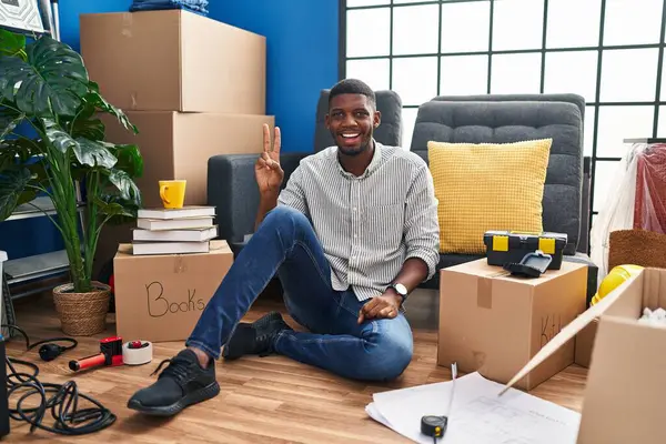 stock image African american man sitting on the floor at new home smiling with happy face winking at the camera doing victory sign with fingers. number two. 