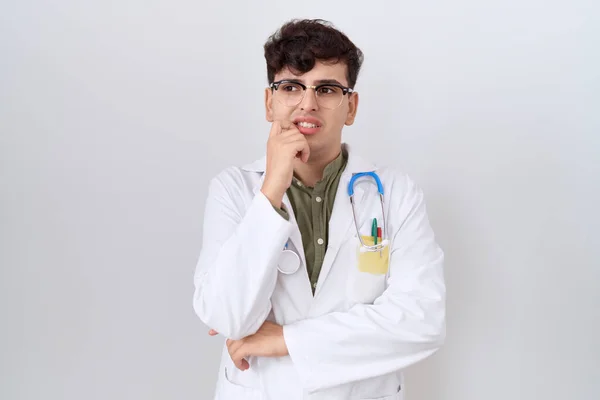 Young non binary man wearing doctor uniform and stethoscope looking stressed and nervous with hands on mouth biting nails. anxiety problem.