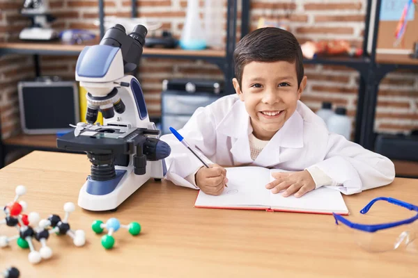 Adorable Hispanic Boy Student Using Microscope Writing Notebook Laboratory Classroom — Stock Photo, Image