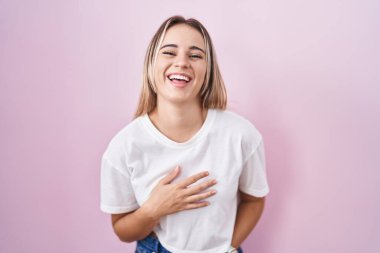 Young blonde woman standing over pink background smiling and laughing hard out loud because funny crazy joke with hands on body. 