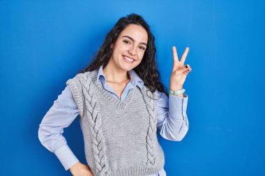 Mavi arka planda duran esmer kadın kameraya bakıp zafer işareti yapıyor. İki numara.. 