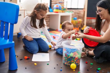 Anaokulunda yerde oturmuş inşaat bloklarıyla oynayan kız ve oğlanlı öğretmen.