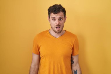 Young hispanic man standing over yellow background in shock face, looking skeptical and sarcastic, surprised with open mouth 