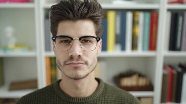 Young hispanic man student sitting on table with relaxed expression at library university