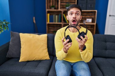 Hispanic man holding broken headphones afraid and shocked with surprise and amazed expression, fear and excited face. 