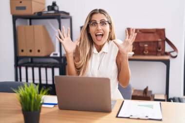 Young blonde woman working at the office wearing glasses celebrating victory with happy smile and winner expression with raised hands 