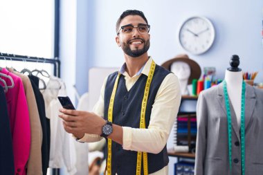 Genç Arap terzi, giyim fabrikasında akıllı telefon kullanarak gülümsüyor.