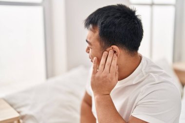 Young chinese man suffering for ear pain sitting on bed at bedroom