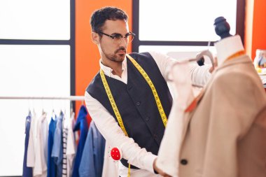 Young hispanic man tailor holding t shirt standing by manikin at atelier
