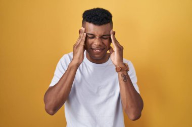 Young hispanic man standing over yellow background with hand on head, headache because stress. suffering migraine. 