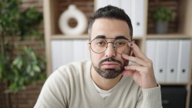 Young hispanic man sitting on sofa with tired expression at office