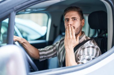 Hispanic man with beard driving car covering mouth with hand, shocked and afraid for mistake. surprised expression 
