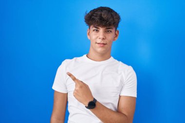 Hispanic teenager standing over blue background pointing aside worried and nervous with forefinger, concerned and surprised expression 