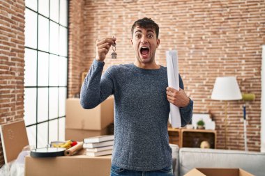 Young hispanic man holding keys of new home and blueprints celebrating crazy and amazed for success with open eyes screaming excited. 