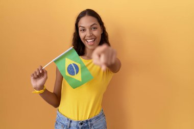 Young hispanic woman holding brazil flag pointing to you and the camera with fingers, smiling positive and cheerful 
