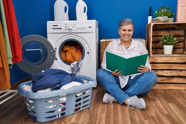 洗濯室で洗濯機を待っている本を読んでいる中年女性 — ストック写真