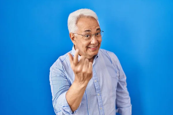 Hispanic senior man wearing glasses beckoning come here gesture with hand inviting welcoming happy and smiling