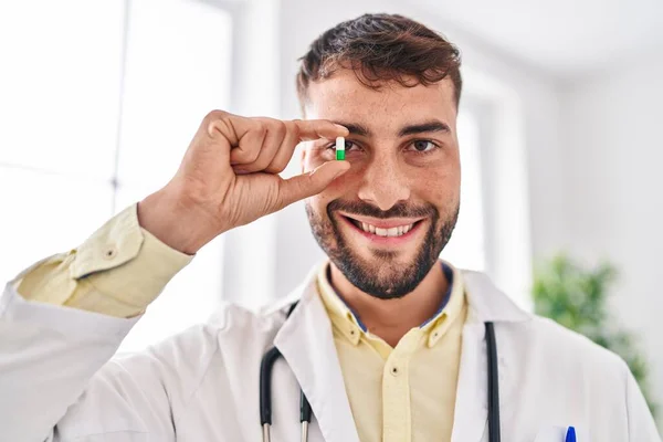 Young hispanic man wearing doctor uniform holding pill over eye at clinic