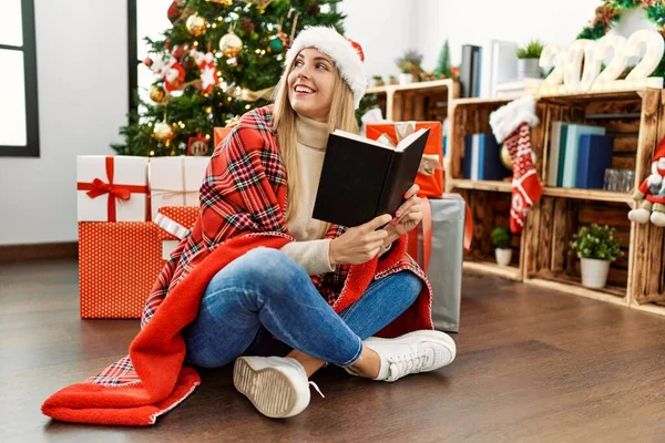 Young blonde woman reading book sitting by christmas tree at home