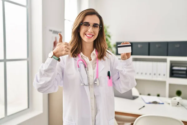 Young doctor woman holding credit card at the clinic smiling happy and positive, thumb up doing excellent and approval sign