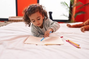 Adorable hispanic girl drawing on notebook lying on sofa at bedroom