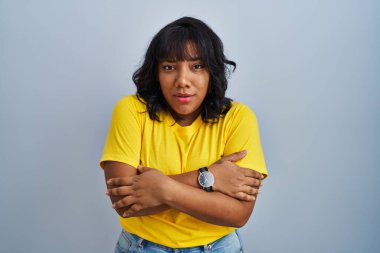 Hispanic woman standing over blue background shaking and freezing for winter cold with sad and shock expression on face 