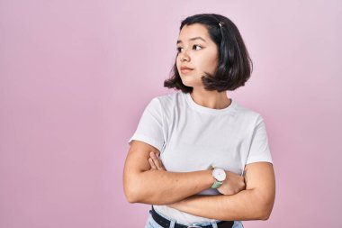 Young hispanic woman wearing casual white t shirt over pink background looking to the side with arms crossed convinced and confident 