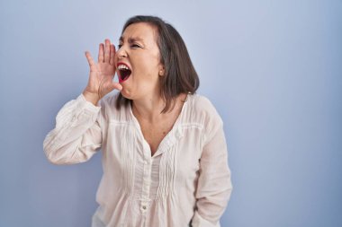 Middle age hispanic woman standing over blue background shouting and screaming loud to side with hand on mouth. communication concept. 