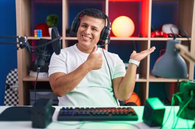 Young hispanic man playing video games showing palm hand and doing ok gesture with thumbs up, smiling happy and cheerful 