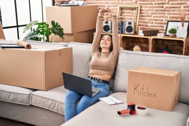 Young beautiful hispanic woman stretching arms using laptop at new home