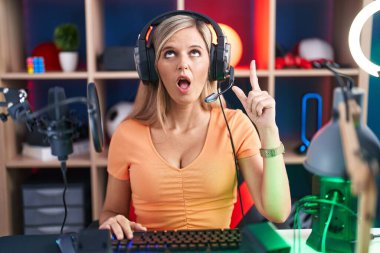 Young woman playing video games amazed and surprised looking up and pointing with fingers and raised arms. 