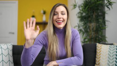 Young blonde woman sitting on sofa saying hello with hand at home