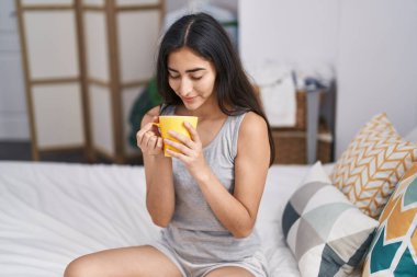 Young hispanic girl drinking cup of coffee sitting on bed at bedroom