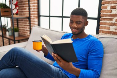 Young african american man reading book and drinking coffee sitting on sofa at home