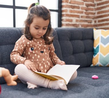 Adorable hispanic girl reading book sitting on sofa at home