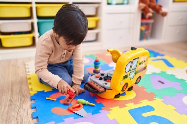 Adorable hispanic boy playing with technician tools toy sitting on floor at kindergarten