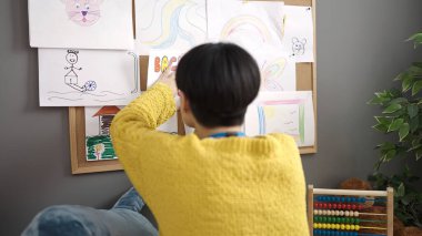 Young chinese woman preschool teacher hanging back to school message on cork board at kindergarten
