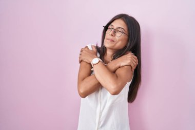 Esmer, pembe arka planda duran, gözlüklü, mutlu, pozitif ve kendinden emin bir şekilde kendine sarılan genç bir kadın. Sevgi ve özgüven. 