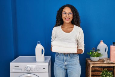 Çamaşırhanede katlanmış havluları tutarken kendinden emin gülümseyen genç Latin kadın.