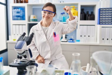 Hispanic girl with down syndrome working at scientist laboratory strong person showing arm muscle, confident and proud of power 